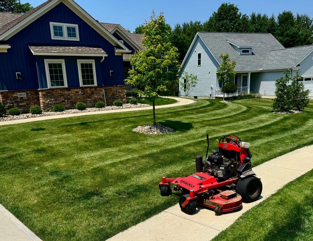 Red lawn mower on a freshly mowed lawn beside two landscaped homes on a sunny day.
