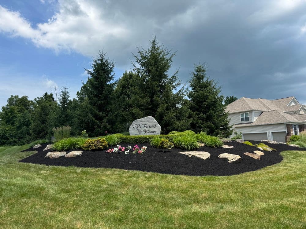 Landscaped entrance to Mill Frant Woods with rocks, flowers, and evergreen trees under cloudy sky.
