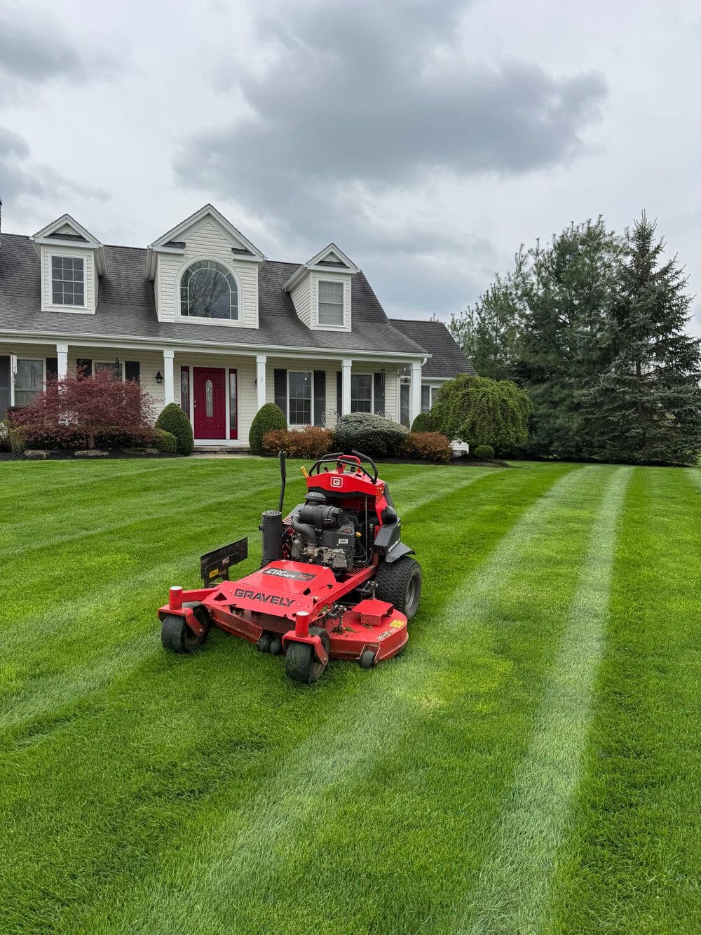 Red Gravely lawn mower cutting stripes on a lush green lawn in front of a suburban home.