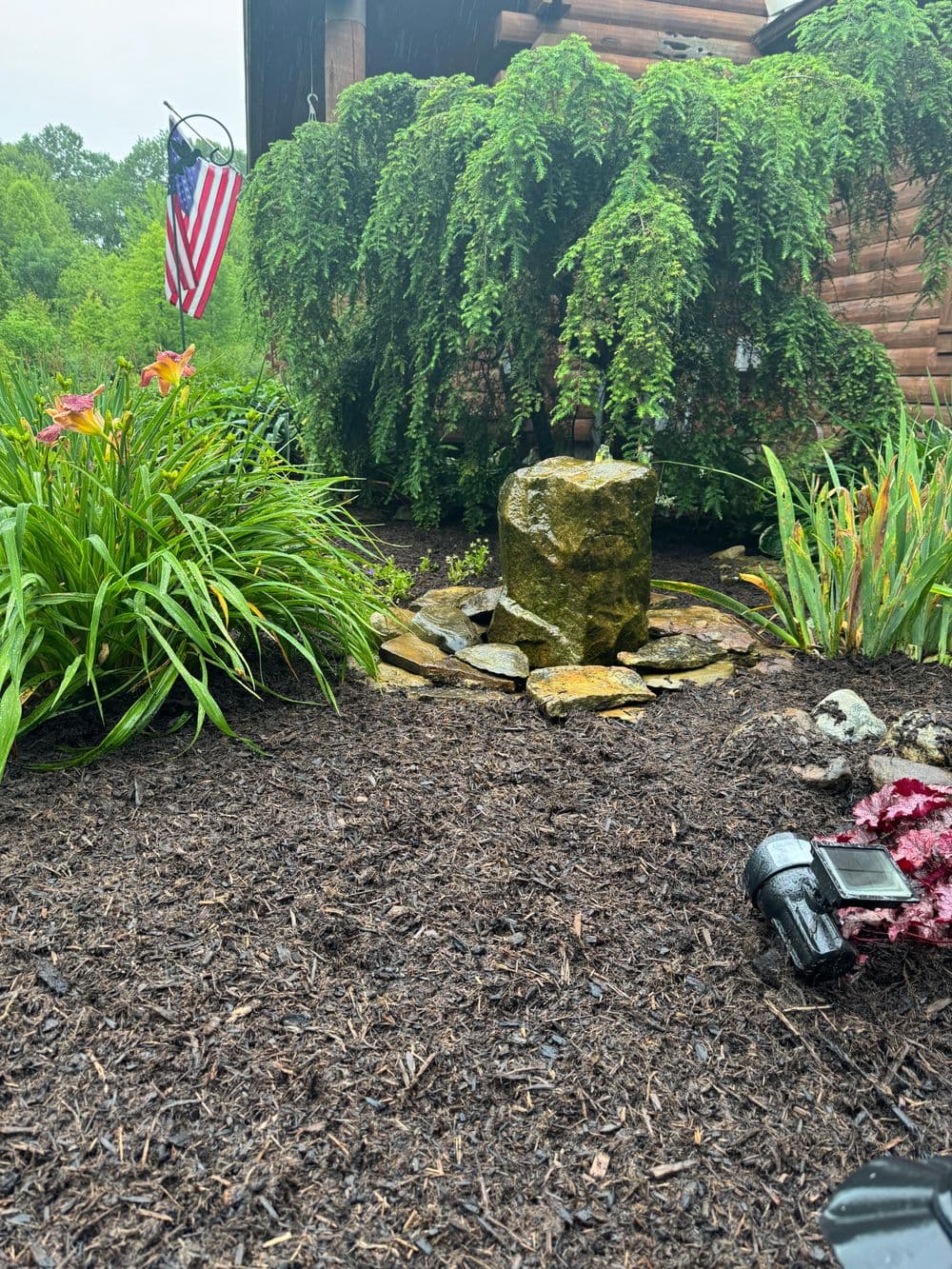 Lush garden featuring a stone fountain, vibrant flowers, and an American flag in the background.