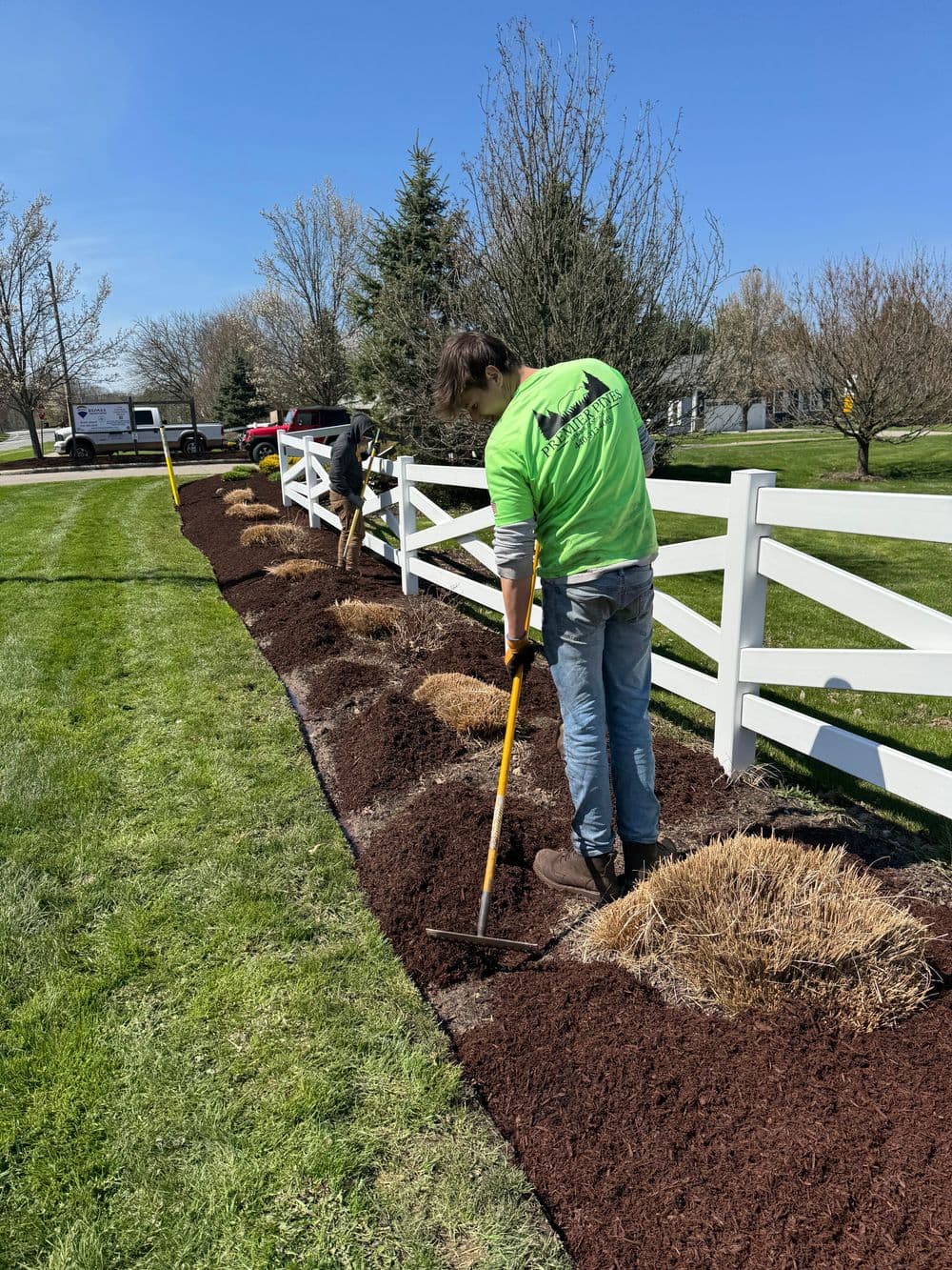 Landscapers applying mulch along a white fence in a sunny outdoor setting.
