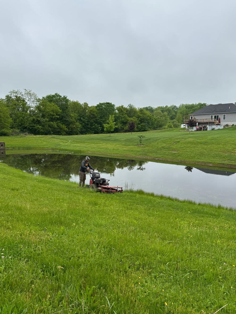 Man mowing grass near a pond with a house and green landscape in the background.