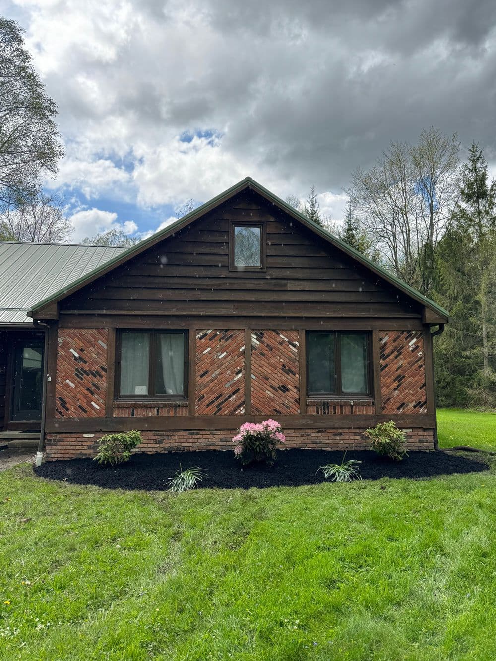 Rustic brick and wood house with flower beds and grassy lawn under a cloudy sky.