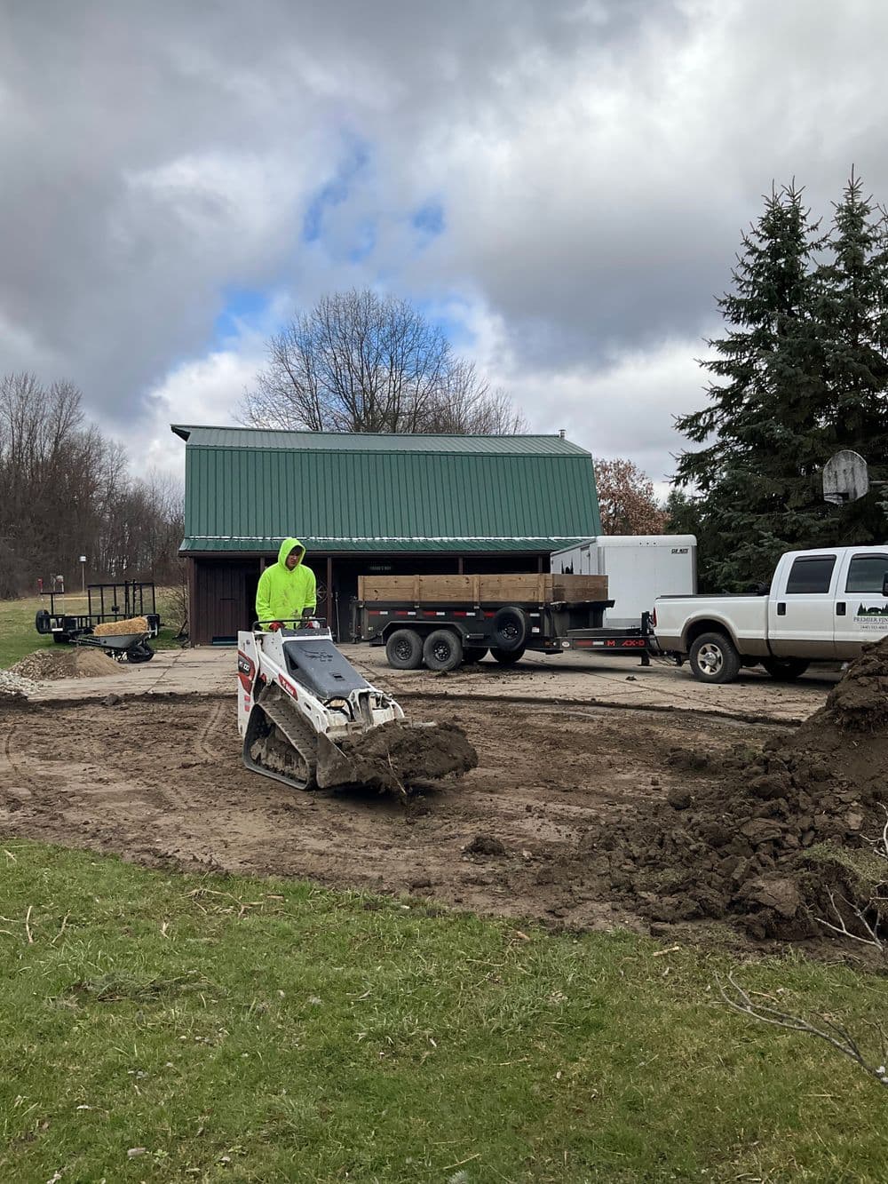 Construction worker using a skid steer loader on a job site with a trailer and barn in background.
