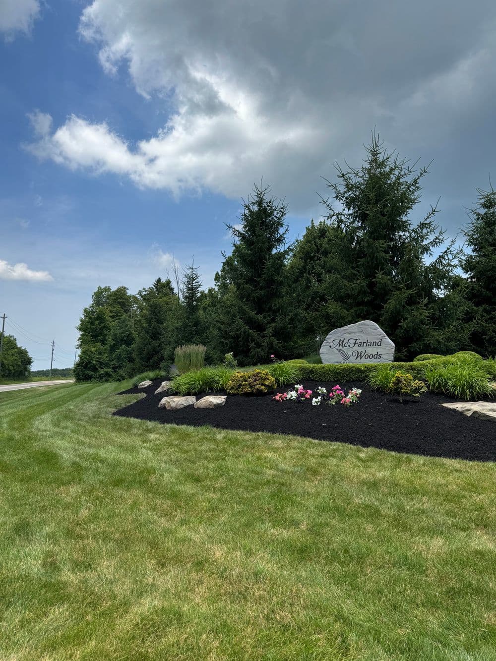 McFarland Woods entrance sign surrounded by lush greenery and colorful flowers under a cloudy sky.