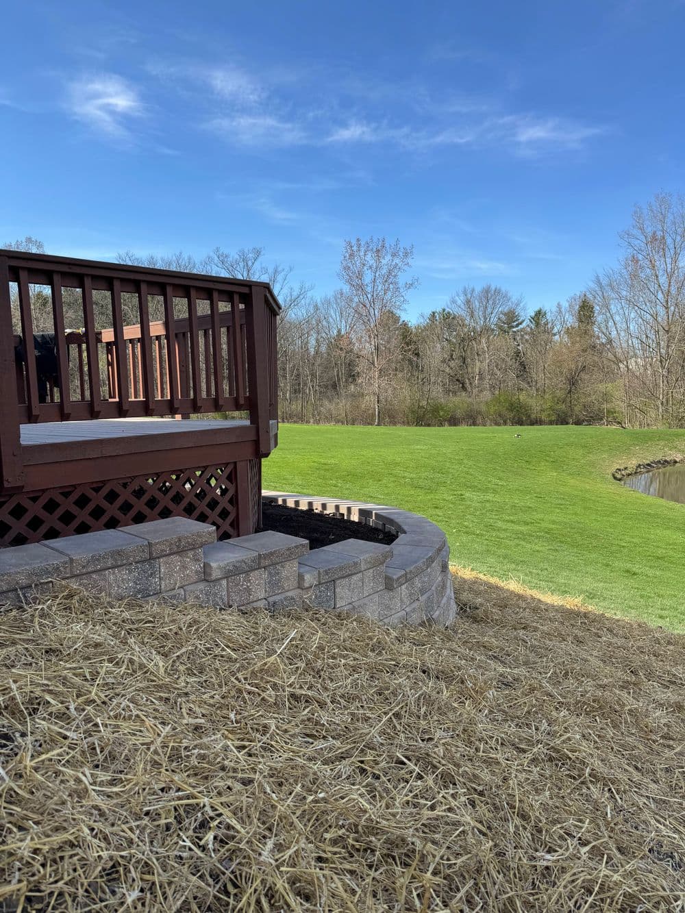 Wooden deck overlooking a green lawn and trees, with a stone retaining wall and blue sky.