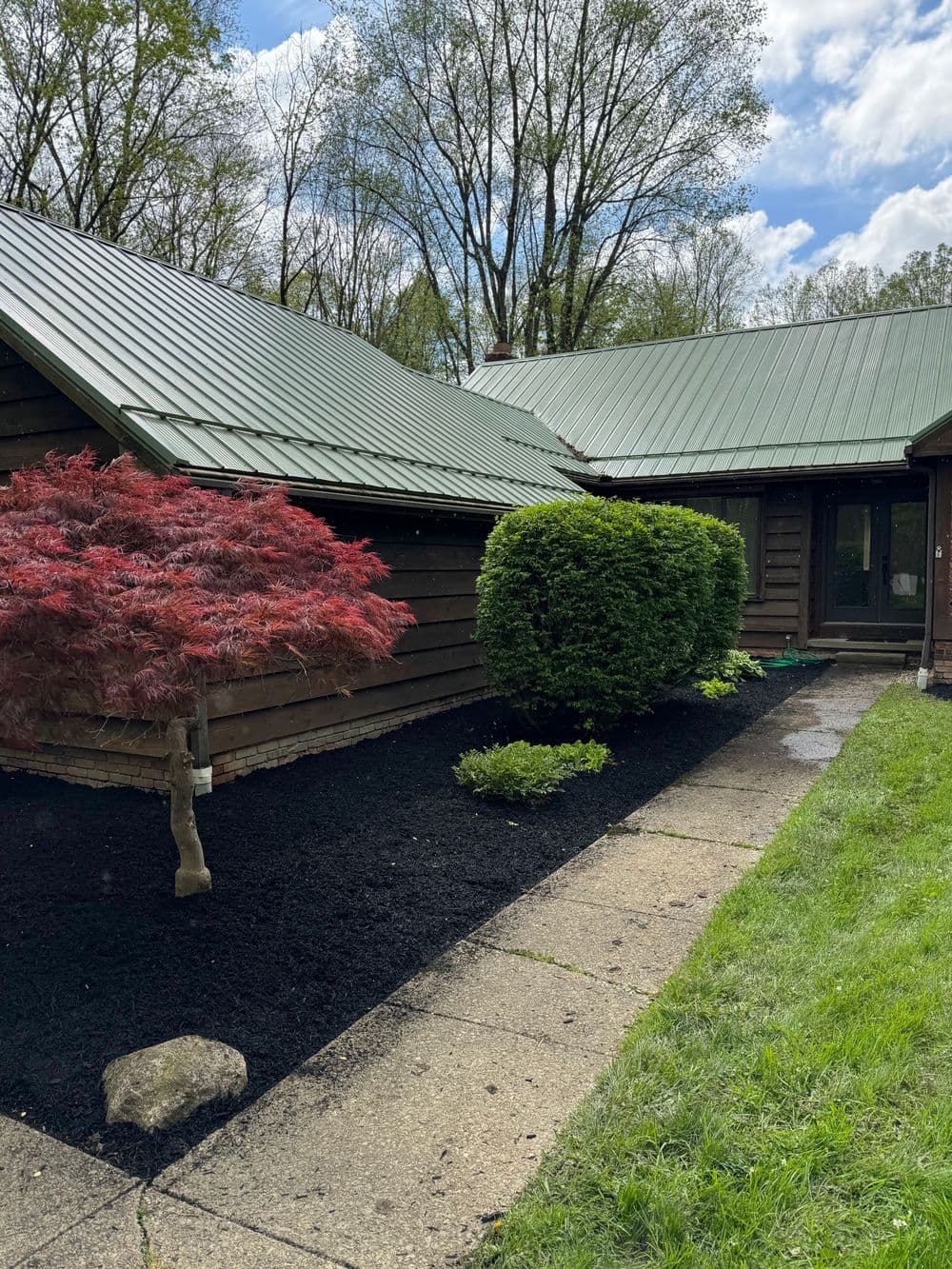 Log cabin exterior with green metal roof, landscaped yard, and blooming Japanese maple tree.