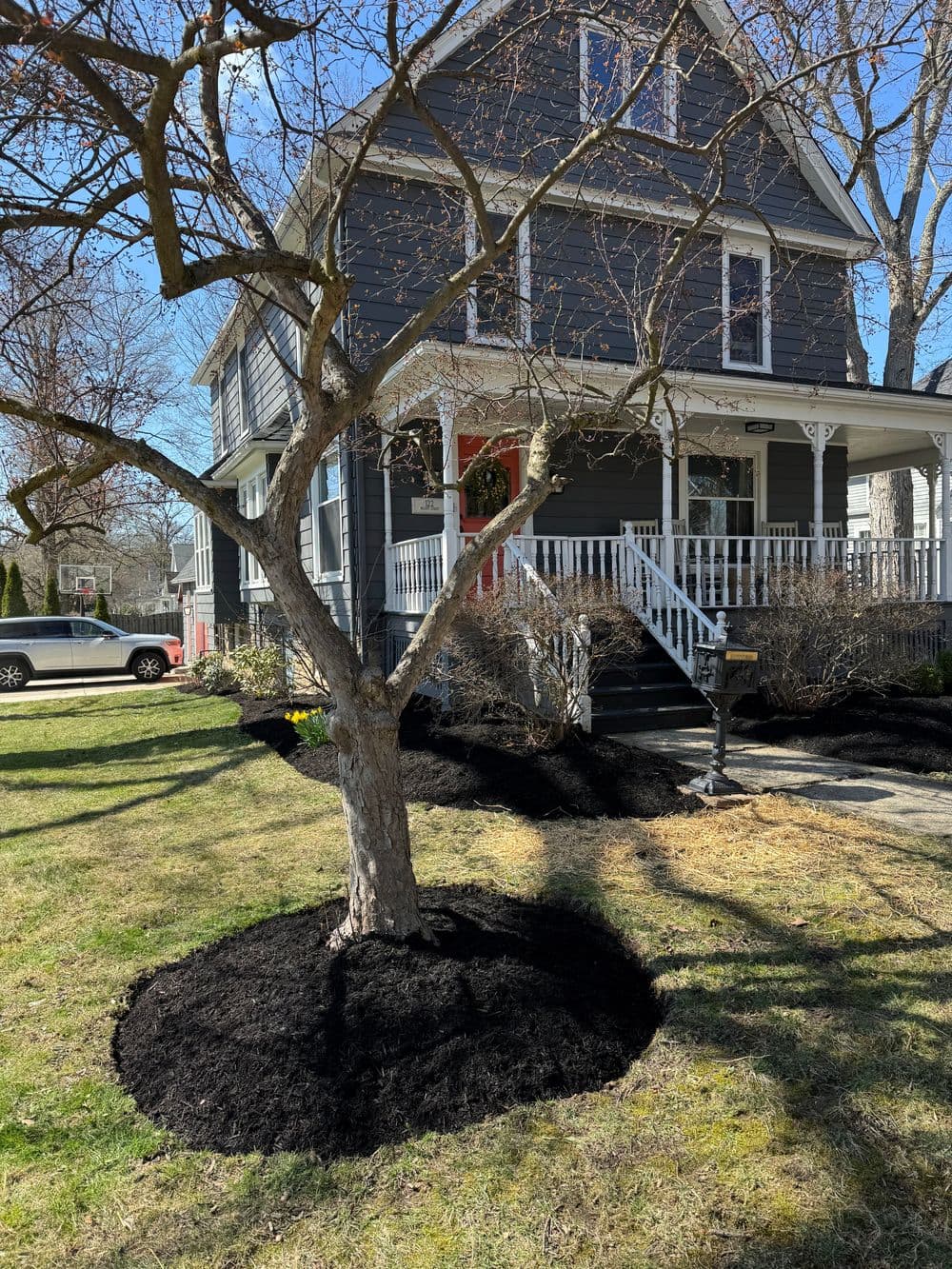 House with gray siding, front porch, newly mulched flower beds, and a tree in spring.