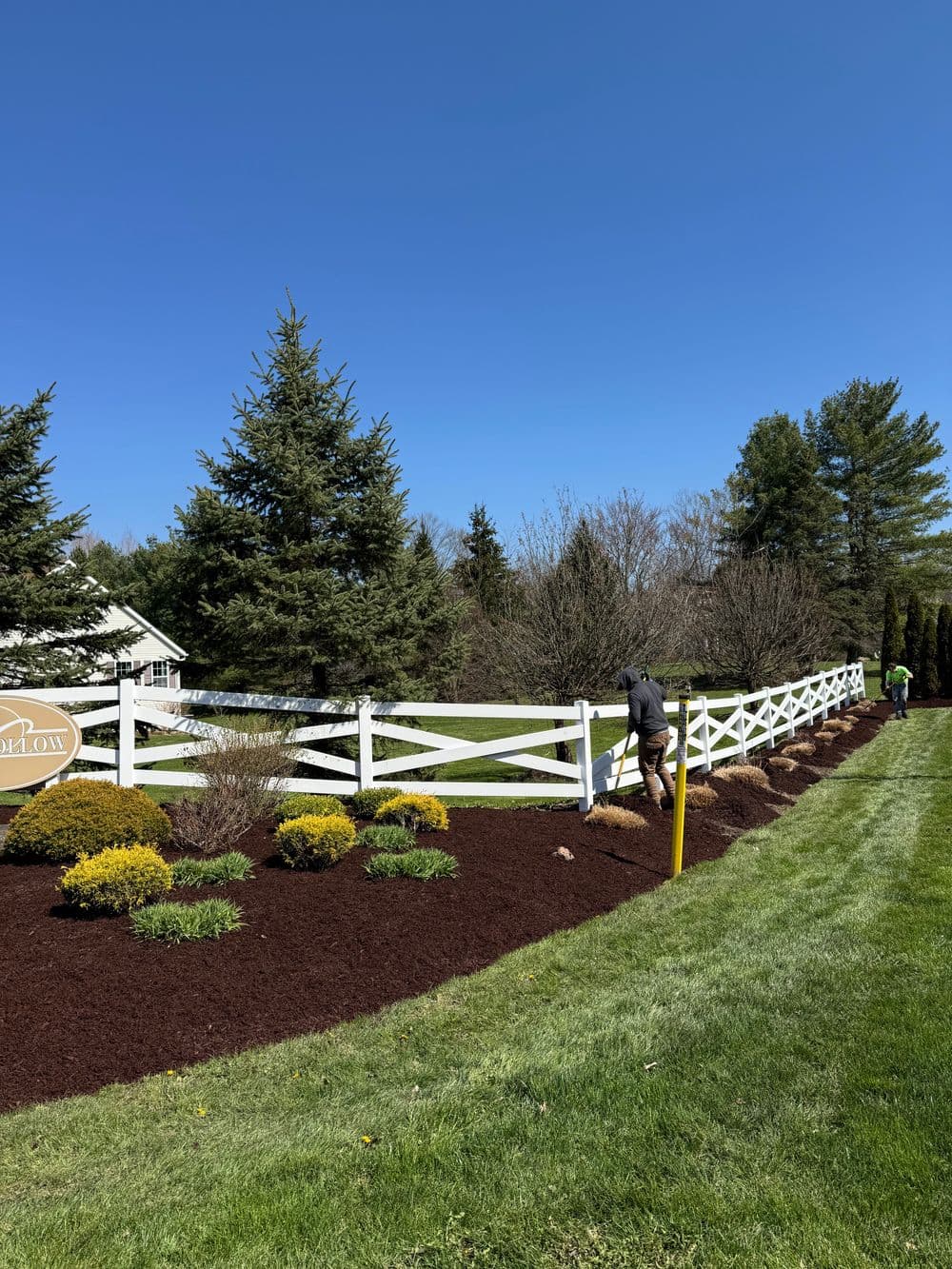 Landscaper tending to flower beds near a white fence on a sunny day. Green grass and trees in background.