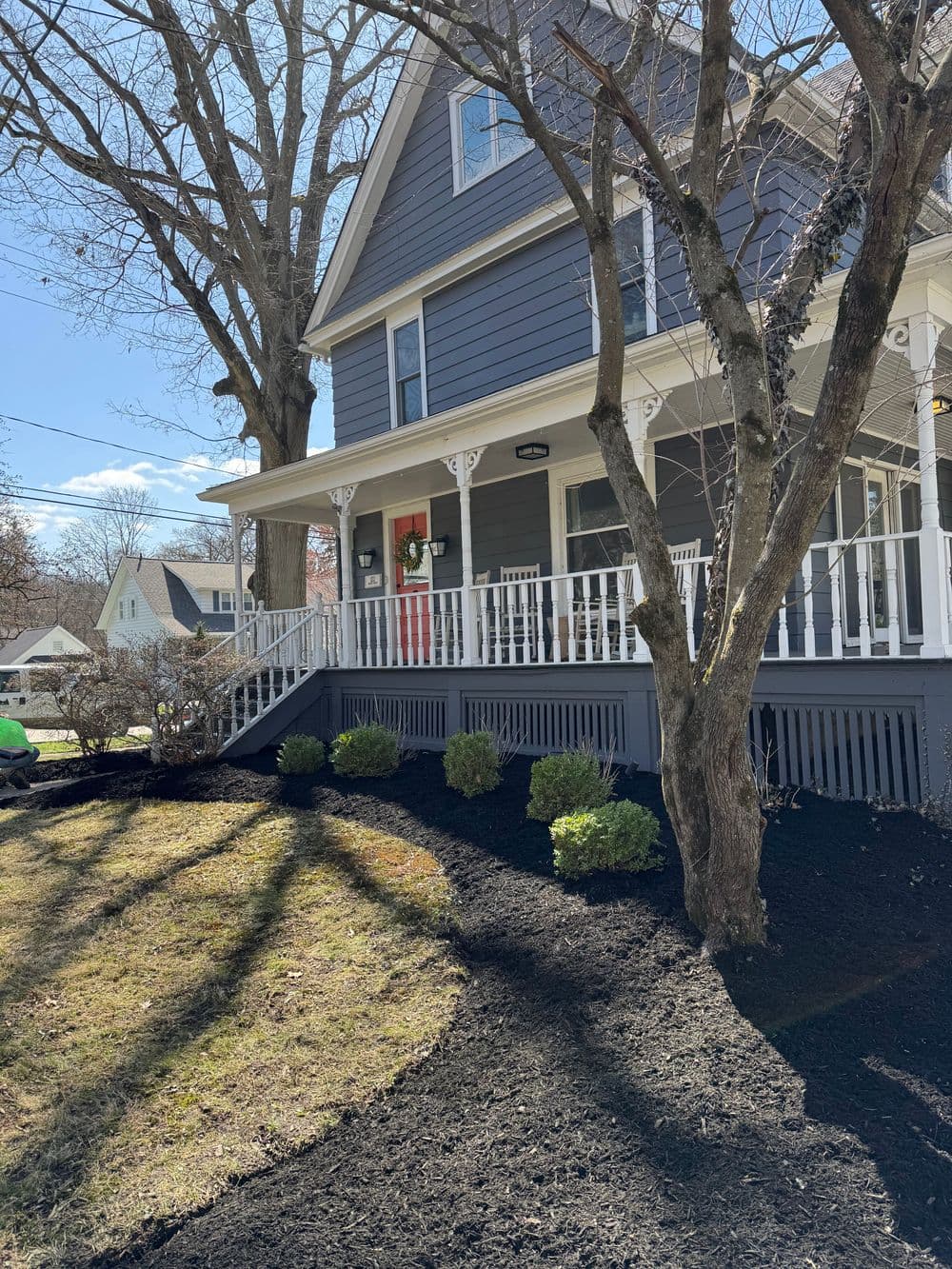 Victorian house with porch, landscaped yard, and freshly mulched garden under sunny sky.