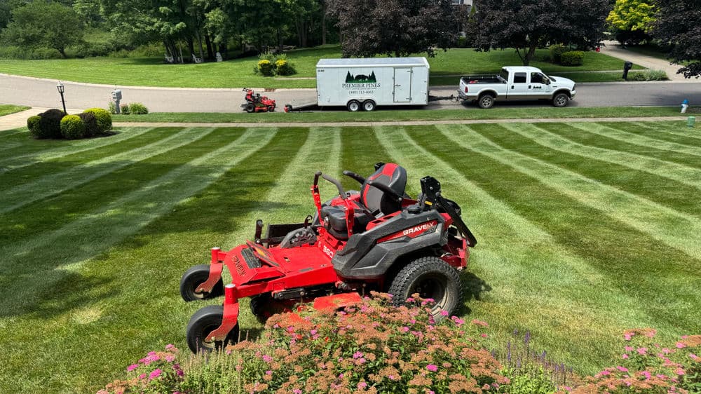 Red riding lawn mower on a striped lawn with a trailer and truck in the background.